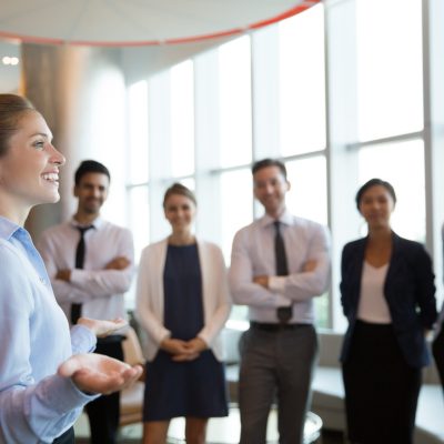 Portrait of successful female executive manager with open hands gesture standing in front of her team in office hall and announce good news