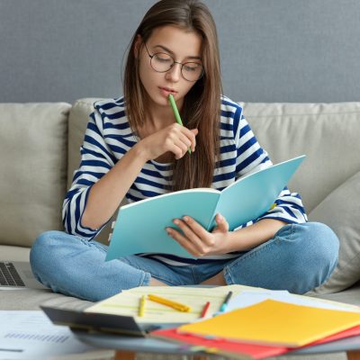 Studying online concept. Serious young woman being busy with remote freelance project, sits at comfortable sofa, writes notes, holds textbook, use laptop computer at home with wireless internet